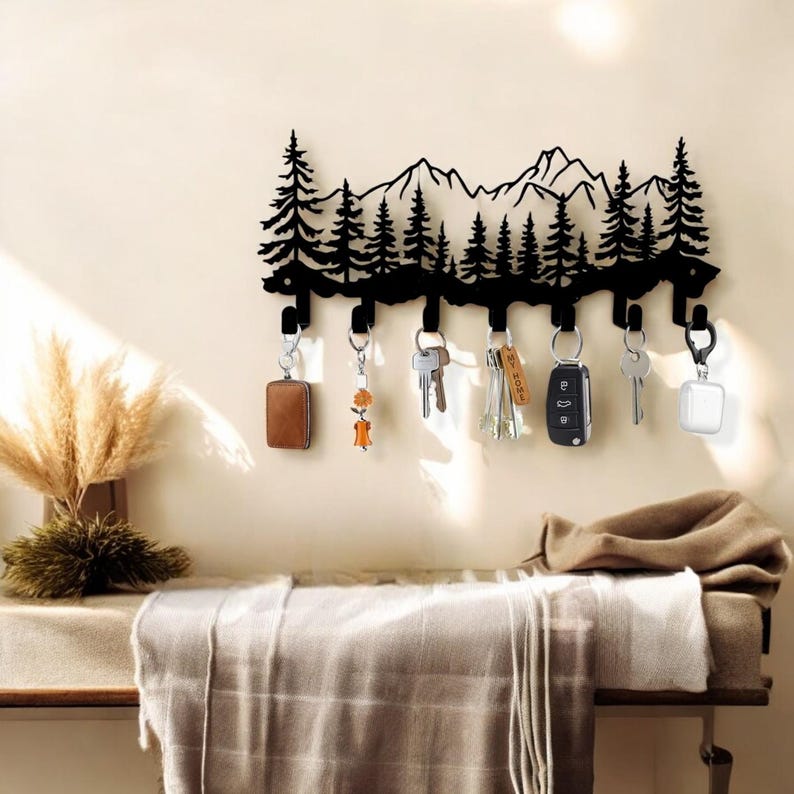 May include: Black metal key holder shaped like a mountain range with pine trees. The holder has six hooks for hanging keys, and there are keys and other items hanging from the hooks.