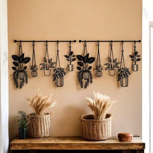 May include: Black metal wall art featuring hanging plant silhouettes. The artwork includes various potted plants suspended from horizontal bars. Below, two woven baskets hold dried ornamental grass, adding a natural touch to the decor. A wooden table and a small wooden bowl complete the scene.