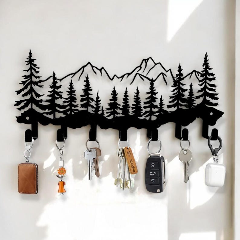 May include: Black metal key holder shaped like a mountain range with pine trees. The holder has 8 hooks and is hanging on a white wall with keys and a car key fob hanging from the hooks. The text "MY HOME" is on one of the key tags.