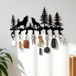 May include: A black metal key holder depicting a forest scene with two howling wolves and pine trees. The key rack has several hooks holding various keys and keychains. The design is mounted on a white wall, contrasting with the dark silhouette.