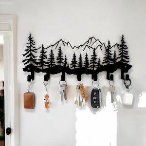 May include: Black metal key holder shaped like a mountain range with pine trees. The holder has six hooks and is mounted on a white wall. Keys and keychains are hanging from the hooks.