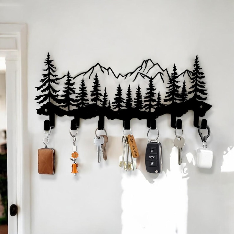 May include: Black metal key holder shaped like a mountain range with pine trees. The holder has six hooks and is mounted on a white wall. Keys and keychains are hanging from the hooks.