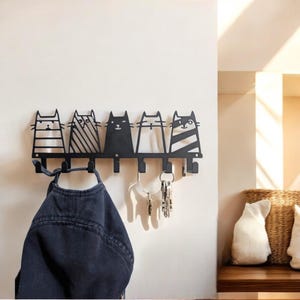 Puede incluir: Perchero y llavero de pared de metal negro con cinco siluetas de gatos estilizadas. Cada gato tiene un diseño único. Una chaqueta vaquera y llaves cuelgan de los ganchos, mostrando la funcionalidad del perchero. El perchero está montado en una pared blanca.