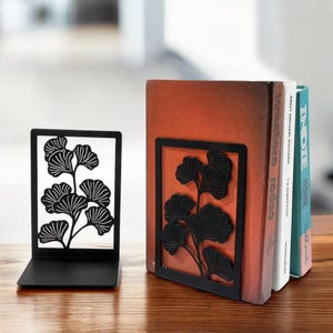 Puede incluir: Un par de sujetalibros de metal negro con un diseño de hoja de ginkgo. Los sujetalibros sostienen dos libros titulados "Indie" y "Western Rodeo".