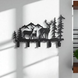 Puede incluir: Perchero de pared de metal negro con una escena de bosque con ciervos, árboles y montañas. El diseño incluye varios ganchos para colgar objetos. El estilo silueta añade un toque rústico a la decoración del hogar.