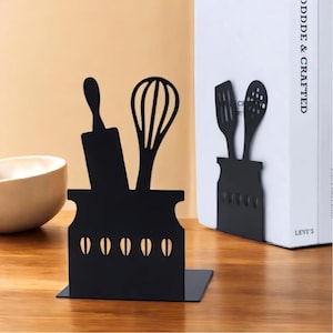 May include: A pair of black metal bookends shaped like jars with kitchen utensils sticking out of the top. The bookends are on a wooden surface.