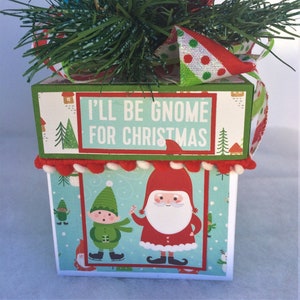 May include: A white gift box with a green and red patterned design. The box has a red and white trim and a green top with the text "I'll be gnome for Christmas". The box features a gnome and elf illustration.