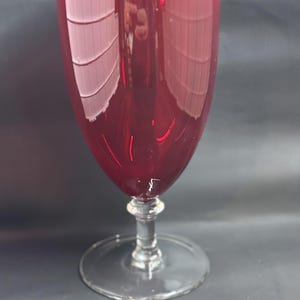 Puede incluir: Una copa de vino alta, de color rojo rubí, con tallo y base transparentes. La copa tiene un cuenco redondeado y un tallo largo y esbelto. La copa está hecha de un material transparente y refleja la luz. La copa está vacía y sobre una superficie oscura.