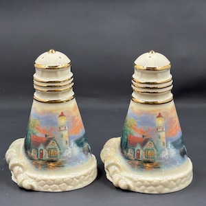 Può includere: Un paio di saliere e pepiere in ceramica. Ogni saliera e pepiera ha la forma di un faro con una scena costiera dettagliata. Le saliere e le pepiere sono color crema con accenti dorati e poggiano su una base a forma di onda. La scena del faro presenta una casa con il tetto rosso e un faro.