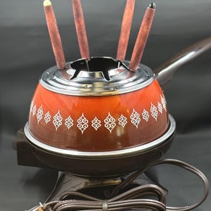 Könnte beinhalten: Ein Vintage-Fondue-Topf mit roter und brauner Emaille-Oberfläche, verziert mit einem dekorativen weißen Muster. Der Topf hat einen silbernen Rand, einen braunen Griff und vier Fonduegabeln mit farbigen Spitzen. Er steht auf einem braunen Sockel mit einem Netzkabel.