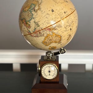 May include: A vintage-style globe with a cream-colored surface, displaying continents and oceans in various colors. The globe is mounted on a wooden base with a built-in barometer. The base is dark brown with brass accents.