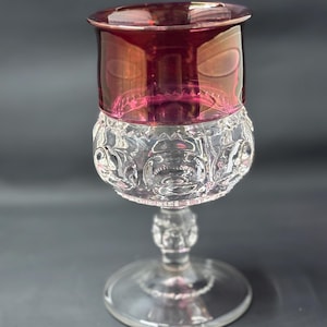 Puede incluir: Una copa de cristal transparente con un borde rojo rubí y un cuenco decorativo con dibujos. La copa tiene un tallo detallado y una base redonda, sobre un fondo oscuro.