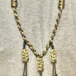 May include: A camouflage-patterned lanyard with three hanging loops. The lanyard is made of braided cord in shades of green, tan, and brown. The loops are knotted at the bottom. The lanyard is designed to be worn around the neck.