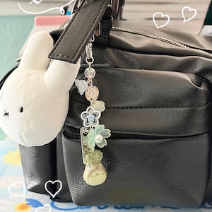 May include: A black leather bag with a white plush bunny charm and a decorative keychain. The keychain features a Totoro figure, floral accents, and clear beads. The word "love" is written in cursive.