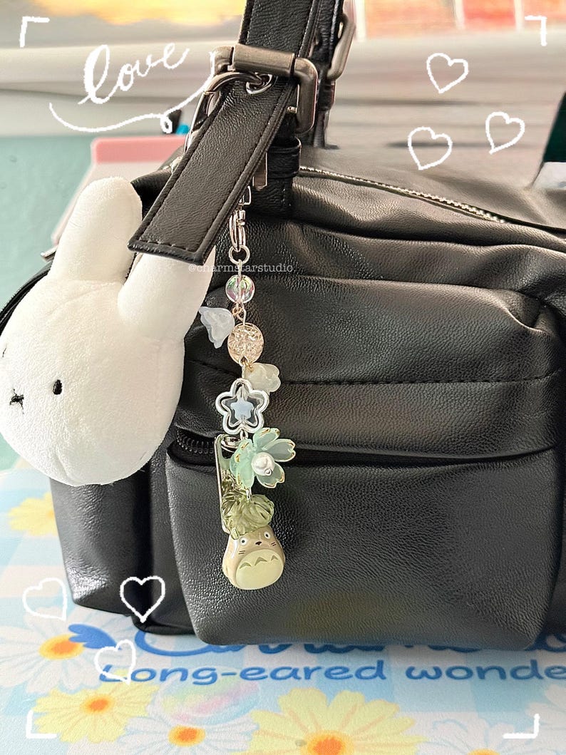 May include: A black leather bag with a white plush bunny charm and a decorative keychain. The keychain features a Totoro figure, floral accents, and clear beads. The word "love" is written in cursive.