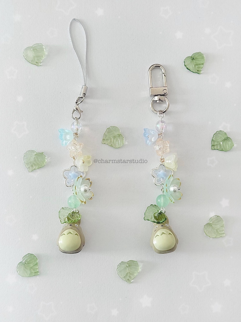 May include: Two beaded charms with a Totoro figure at the base. The charms feature light blue and white flower beads, green leaf beads, and clear beads. One charm has a silver lanyard, and the other has a silver clasp. The text "@charmstarstudio" is visible.
