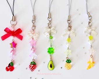 May include: Five colorful phone charms with fruit and bow accents. The charms feature a red cherry, a strawberry, an avocado, a pineapple, and a banana. Each charm has a white bow and a string for attaching to a phone.