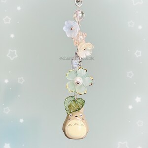 May include: A whimsical keychain featuring a Totoro figure, adorned with delicate flower and leaf charms. The keychain includes a silver-tone clasp and is accented with clear and pastel-colored beads. The text "@charmstarstudio" is visible.