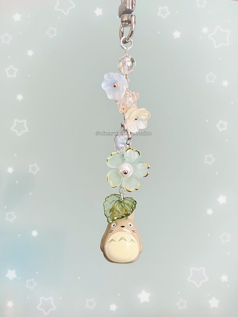 May include: A whimsical keychain featuring a Totoro figure, adorned with delicate flower and leaf charms. The keychain includes a silver-tone clasp and is accented with clear and pastel-colored beads. The text "@charmstarstudio" is visible.