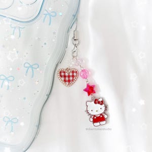 May include: A phone charm with a Hello Kitty pendant in a red dress, a red gingham heart, pink beads, and red star charms. The charm is attached to a clear phone case with bow and star designs.