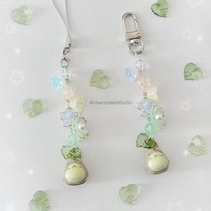 May include: Two beaded phone charms with a cartoon character, flowers, and beads in pastel colors. One charm has a silver clasp, the other a silver loop. Green leaf-shaped embellishments and the text "@charmstarstudio" are scattered around the charms.