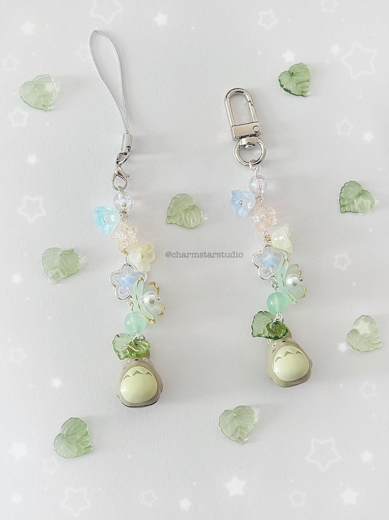 May include: Two beaded phone charms with a cartoon character, flowers, and beads in pastel colors. One charm has a silver clasp, the other a silver loop. Green leaf-shaped embellishments and the text "@charmstarstudio" are scattered around the charms.