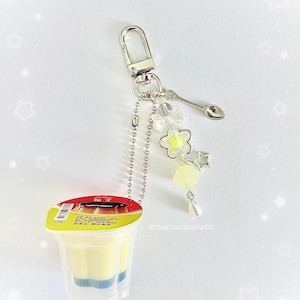May include: A clear plastic keychain charm featuring a small dessert cup with a yellow and blue layered filling. The charm includes a silver spoon, flower and star accents, and a lobster clasp.
