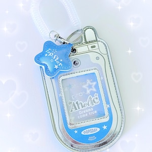 May include: Silver and blue flip phone-shaped keychain with a star-shaped charm. The charm reads "Top Star" and the phone screen displays the text "AhoAc Dreams come true" and "OOPSDAY". The keychain has a clear, coiled wrist strap.