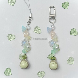 May include: Two beaded charms with a Totoro figure at the bottom. The charms feature light blue and white flower beads, green leaf beads, and clear beads. The charms have a silver clasp and a silver cord.