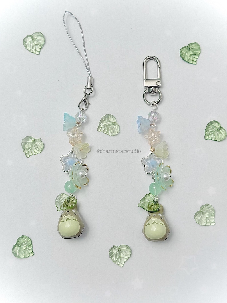 May include: Two beaded charms with a Totoro figure at the bottom. The charms feature light blue and white flower beads, green leaf beads, and clear beads. The charms have a silver clasp and a silver cord.