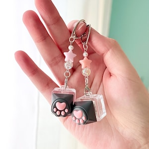 May include: A pair of dangle earrings featuring clear, cube-shaped charms with black paw prints and pink heart-shaped pads. Each earring has a pink star, clear beads, and a pearl. The earrings are suspended from silver-colored ear wires.