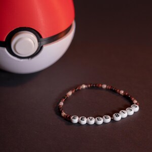 May include: A red and white Poké Ball is positioned near a beaded bracelet. The bracelet spells "EEVEE #133" with white letter beads, interspersed with brown and white beads. The background is a dark, plain colour.