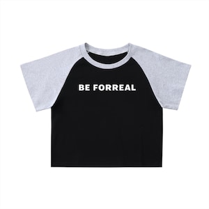 Puede incluir: Top corto raglán negro y gris con el texto "BE FORREAL" en blanco. La camiseta tiene cuello redondo y mangas cortas. Las mangas son grises y el cuerpo de la camiseta es negro.