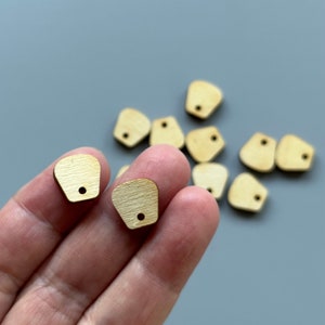 May include: A close-up of a hand holding two small, unfinished wooden shapes with a hole in the top. The shapes are light brown and have a slightly textured surface. There are several other similar shapes scattered on a grey surface.