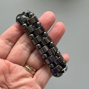 May include: A black hematite bracelet with round beads and cylindrical beads. The bracelet is made of multiple strands of beads.