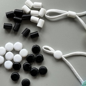 May include: Assortment of black and white plastic mask strap adjusters and stoppers. The adjusters are cylindrical and the stoppers are round.