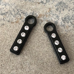 May include: Two black metal keychains with a loop at the top and five clear rhinestones on the front.