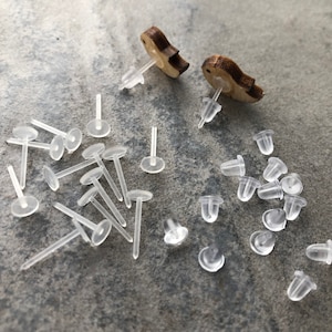 May include: A close-up of a pile of clear plastic earring backs. There are two wooden earring posts with a bird design in the background.