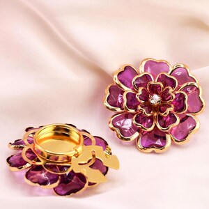 May include: Two decorative flower-shaped candle holders. The petals are a deep purple with gold trim. One holder has a gold interior for a candle, and the other has a gold centre with a small crystal. The background is a soft, pink fabric.