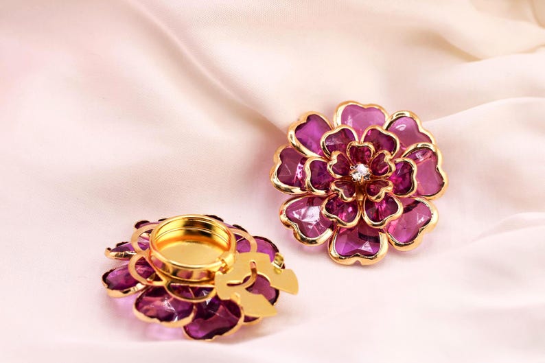May include: Two decorative flower-shaped candle holders. The petals are a deep purple with gold trim. One holder has a gold interior for a candle, and the other has a gold centre with a small crystal. The background is a soft, pink fabric.