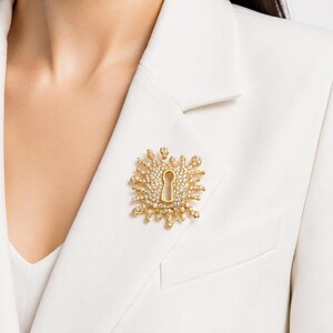 May include: A gold-toned brooch in the shape of a keyhole, encrusted with clear crystals, is pinned to a white blazer. The brooch has a sunburst design around the keyhole. The blazer is worn by a person.