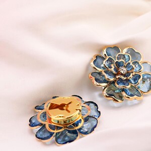 May include: Two flower-shaped accessories with blue and gold accents. The petals are a translucent blue, and the centre of one has a small, sparkling gem. The other has a gold centre. The accessories are on a white, silky surface.