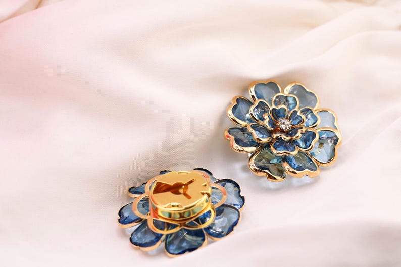 May include: Two flower-shaped accessories with blue and gold accents. The petals are a translucent blue, and the centre of one has a small, sparkling gem. The other has a gold centre. The accessories are on a white, silky surface.