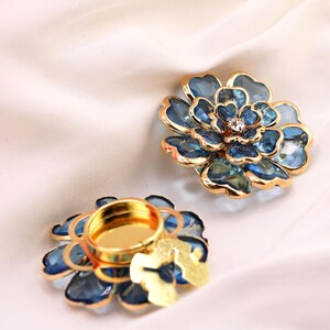 May include: Two flower-shaped candle holders with a blue and gold design. The petals are a gradient of blue, and the edges are outlined in gold. One candle holder has a small, round, gold-coloured cup in the centre.