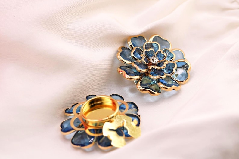 May include: Two flower-shaped candle holders with a blue and gold design. The petals are a gradient of blue, and the edges are outlined in gold. One candle holder has a small, round, gold-coloured cup in the centre.