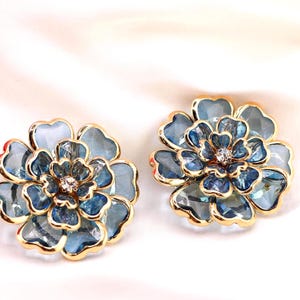 Op de afbeelding: Een paar bloemvormige oorstekers. Elke oorbel heeft een blauw en goud ontwerp met een centrale kristal. De bloemblaadjes zijn lichtblauw en de randen zijn goudkleurig omlijnd. De oorbellen zijn geplaatst tegen een witte achtergrond.