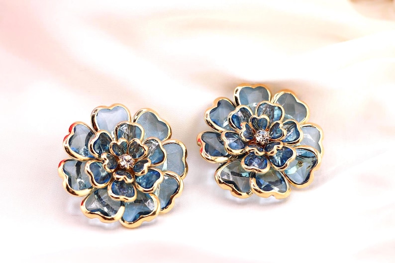May include: A pair of flower-shaped stud earrings. Each earring has a blue and gold design with a central crystal. The petals are a light blue colour, and the edges are outlined in gold. The earrings are set against a white background.