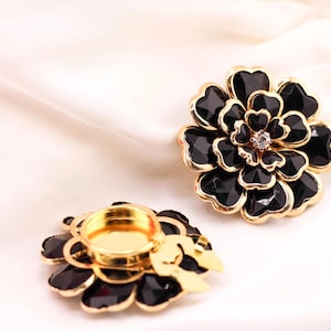 May include: Two black and gold flower-shaped candle holders. The petals are black with a faceted design, and the edges are gold. One holder has a gold-coloured interior for a candle, and the other has a crystal in the centre. The background is a white, silky fabric.