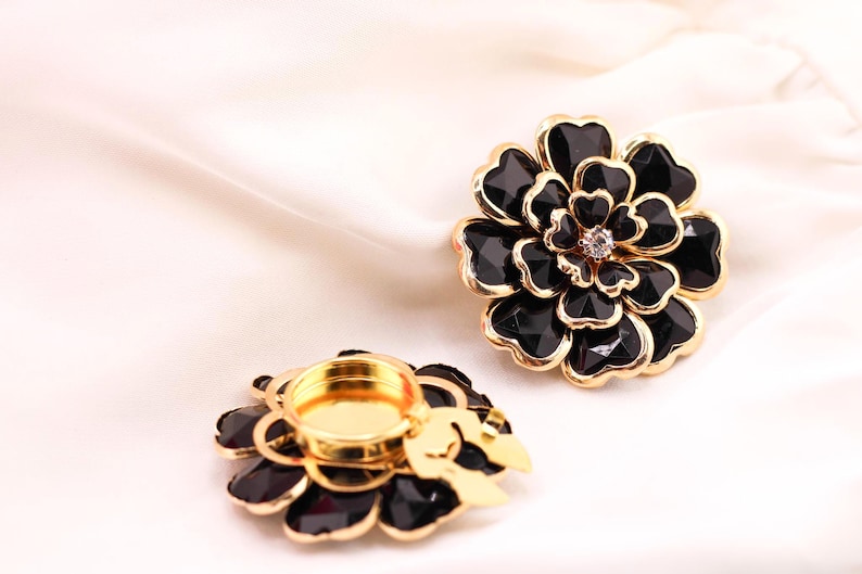 May include: Two black and gold flower-shaped candle holders. The petals are black with a faceted design, and the edges are gold. One holder has a gold-coloured interior for a candle, and the other has a crystal in the centre. The background is a white, silky fabric.