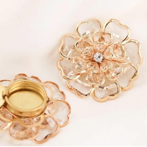 May include: Two flower-shaped decorative objects. The flowers have a gold-coloured metal frame with clear, faceted petals. One flower has a small, round, gold-coloured base. The other flower has a small, clear, round gem in the centre.